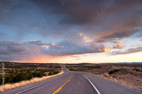 Highway curving into the distance through the landscape near Santa Fe,  New Mexico underneath a dramatic colorful sky at sunset