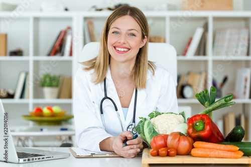 Beautiful smiling nutritionist looking at camera and showing healthy vegetables in the consultation.