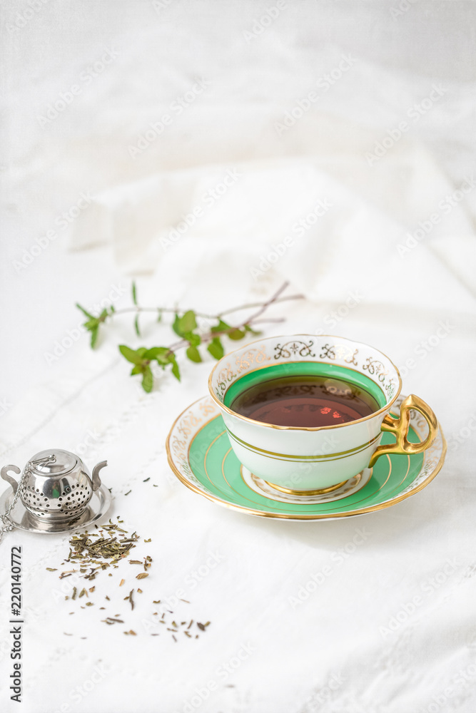 Royal Stafford Fine Bone China Tea Cup and Saucer with Loose Leaf Mint Tea, Spring of Fresh Mint in Background, and Tea Infuser on White Background Royal Stafford Fine Bone China Tea Cup and Saucer with Loose Leaf Mint Tea, Spring of Fresh Mint in Background, and Tea Infuser on White Background