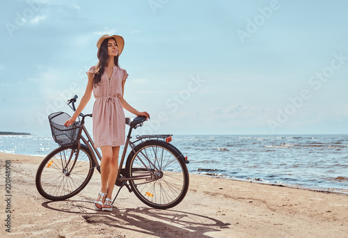 Beautiful brunette girl dressed in dress and hat posing with a bicycle on the beach on a sunny day.