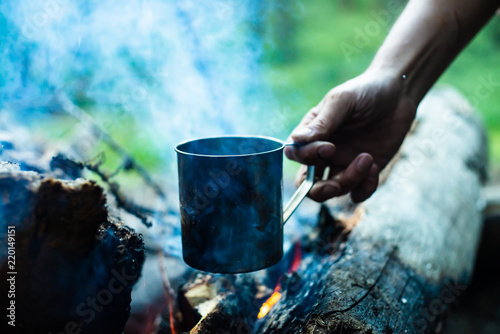 Man is heating tea in metal mug on bonfire. Hot drink on nature. Tea drinking in open air. Steel mug in human hand. Camping in dusk. Romantic warm atmosphere outdoor in twilight . Active rest.