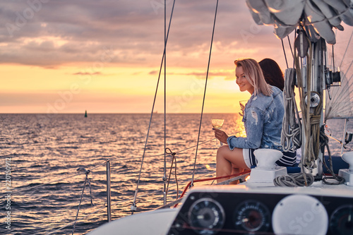 Fototapeta Naklejka Na Ścianę i Meble -  Female friends relaxing on the yacht with glasses of wine in the hands, during sunset on the high seas.