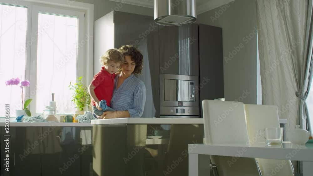 Young woman with curly hair holding little son and teaching him to wipe kitchen surfaces with cloth Young woman with curly hair holding little son and teaching him to wipe kitchen surfaces with cloth