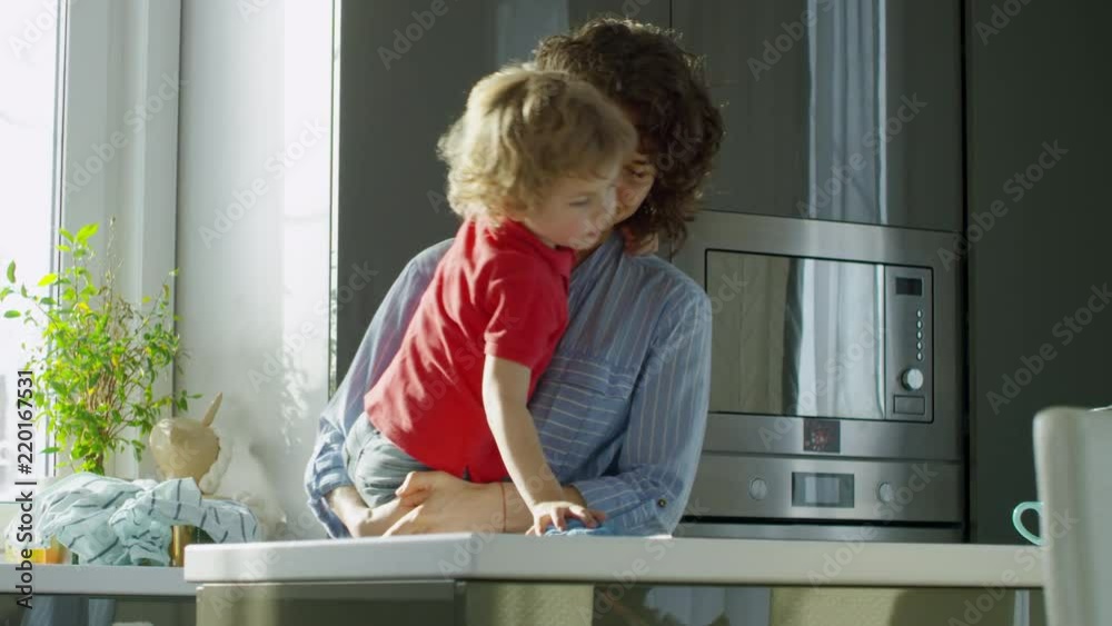 Medium shot of young curly haired mother holding little son and smiling while he is trying to wipe kitchen worktop Medium shot of young curly haired mother holding little son and smiling while he is trying to wipe kitchen worktop