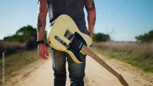 Cool musician walking down dusty track with guitar.