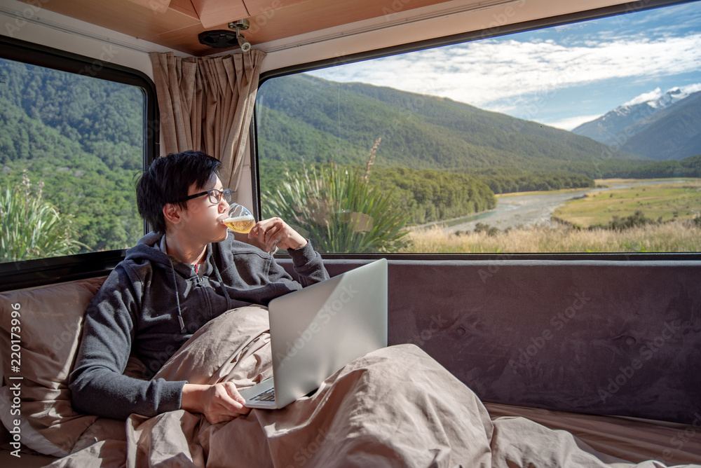 Fototapeta premium Young Asian man drinking beer and working with laptop computer on the bed in camper van with mountain scenic view through the window, digital nomad concept