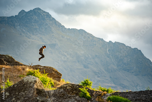 Young man jumping on Queenstown hill with mountain scenery in the background during golden hour sunset in Queenstown, South Island, New Zealand. Enjoy travelling concept