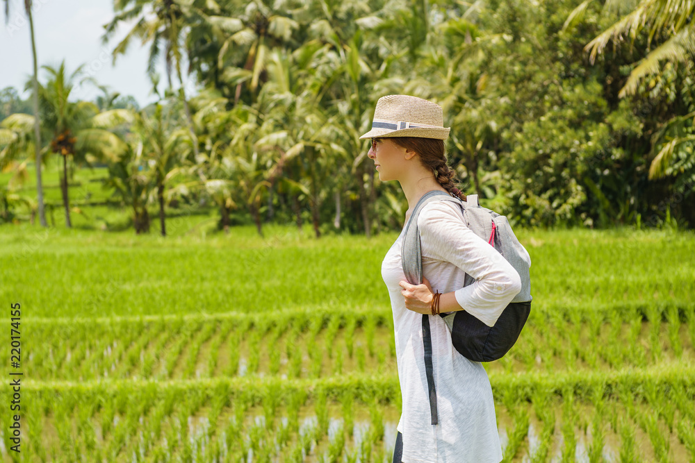 Beautiful young girl traveler in hat, glasses, with backpack walks along a rice field with green grass, palms, amazing blue sky with white clouds on the background has journey in Bali island Indonesia