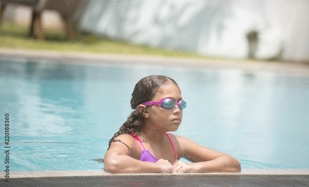 Young child  wearing goggles relaxing at edge of the pool after swimming and playing showing her wet curly hair