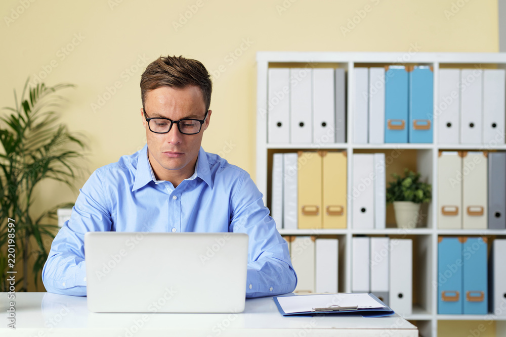 Handsome young man in glasses working on laptop at his table in office Handsome young man in glasses working on laptop at his table in office