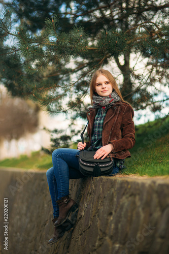 A beautiful girl is walking along the embankment. Blond hair and brown jacket. Spring