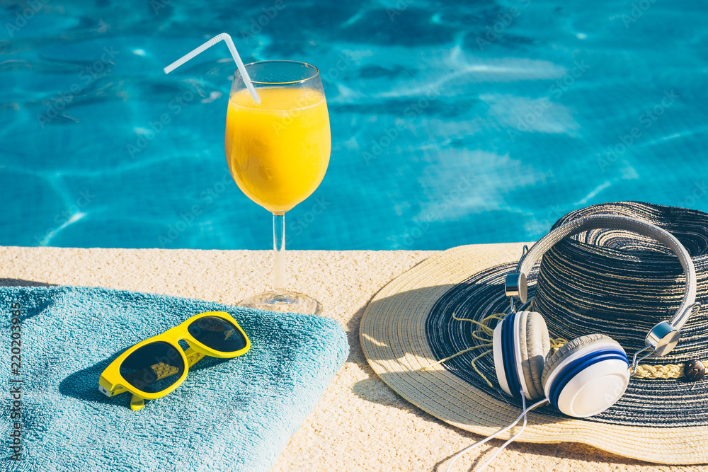 A set of things to relax by the pool on vacation - sunglasses, a hat, a glass of orange juice, headphones