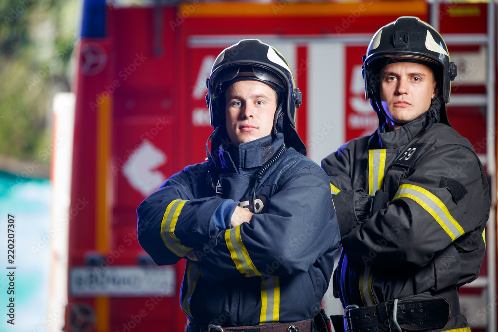 Photo of two firemen with hands on waist near fire engine Photo of two firemen with hands on waist near fire engine