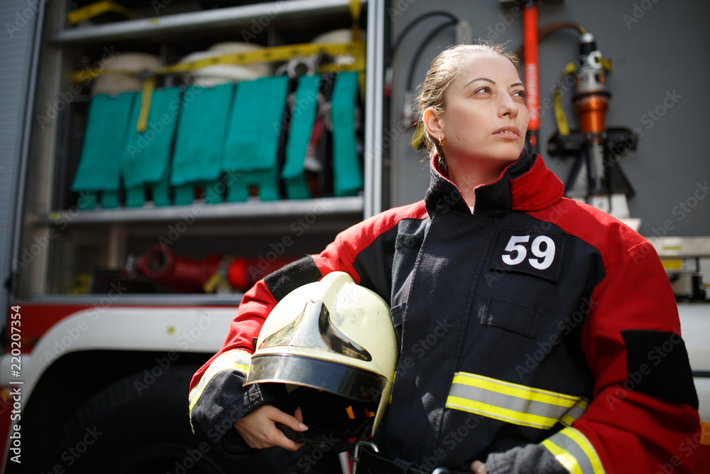 Photo of young woman firefighter looking to side next to fire engine Photo of young woman firefighter looking to side next to fire engine
