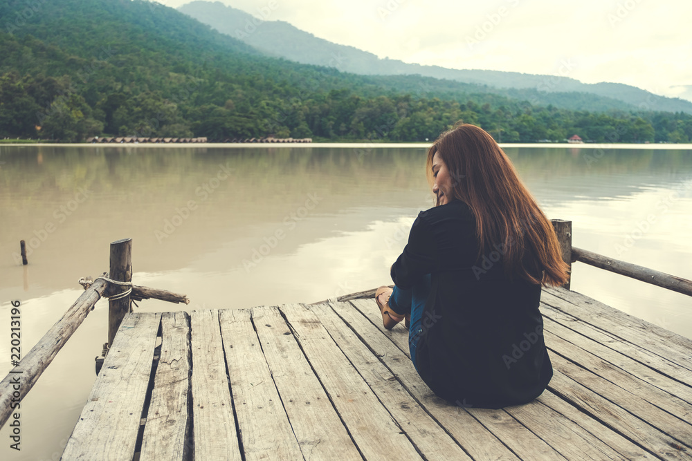 Closeup image of an asian woman sitting alone on an old wooden pier by the river with sky and mountain background