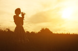 Young woman in dress with bouquet of flowers in hands at sunset in the field. Tinted warm silhouette image