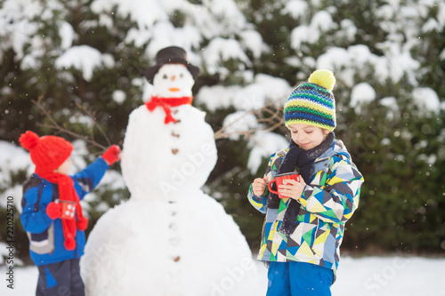 Happy beautiful children, brothers, building snowman in garden and drinking tea