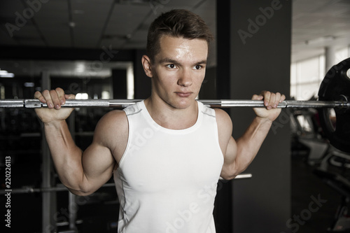 Confident muscular man trainings squats with barbells overhead in gym