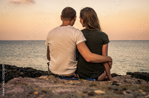 Man and woman hugging sitting on beach and watching the calm sea at sunset