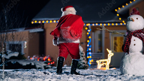 Authentic Santa Claus Tosses Red Bag Over His Shoulder and Walks into Front Yard of the Idyllic House Decorated with Lights and Garlands. Magical New Year's Eve with Falling Snow.