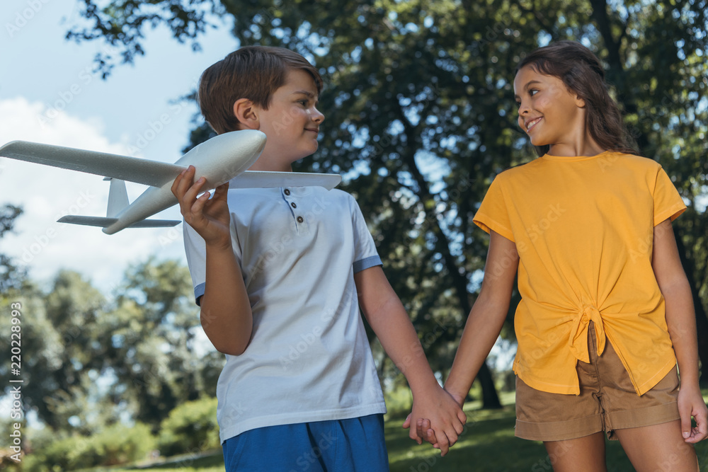 low angle view of happy children holding plane model and smiling each other while holding hands and walking together in park low angle view of happy children holding plane model and smiling each other while holding hands and walking together in park