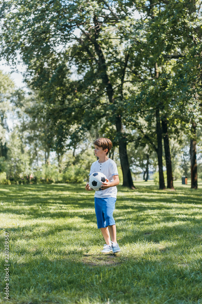 cute little boy holding soccer ball and looking away in park