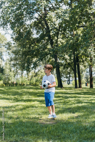 cute little boy holding soccer ball and looking away in park