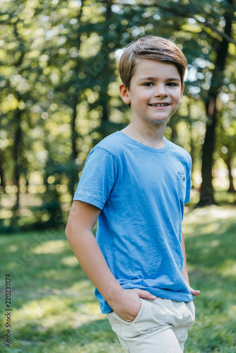 portrait of adorable child standing with hands in pockets and smiling at camera in park