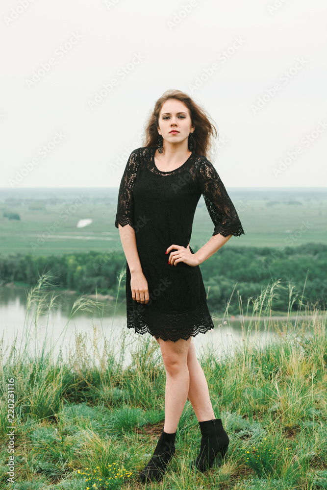 Cheerful young girl in black lacy dress is dancing in meadow on hill above river. Emotion vintage female portrait on nature in sunny day. Recreation on open air. Cheerful young girl in black lacy dress is dancing in meadow on hill above river. Emotion vintage female portrait on nature in sunny day. Recreation on open air.