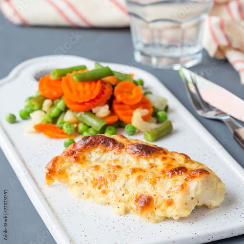 Baked cod fish fillet under cheese, mustard, pepper and cream crust, served with steamed vegetables, on ceramic board, square format
