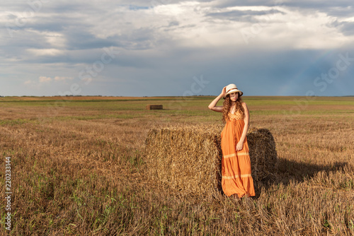 curly young rural teenager girl stands near a bundle of hay in a sundress and hat on a harvested wheat field with a backdrop of a sunset rain sky and a rainbow