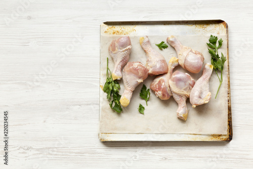 Uncooked raw chicken legs on a baking sheet paper on tray, high angle view. White wooden background. Copy space.