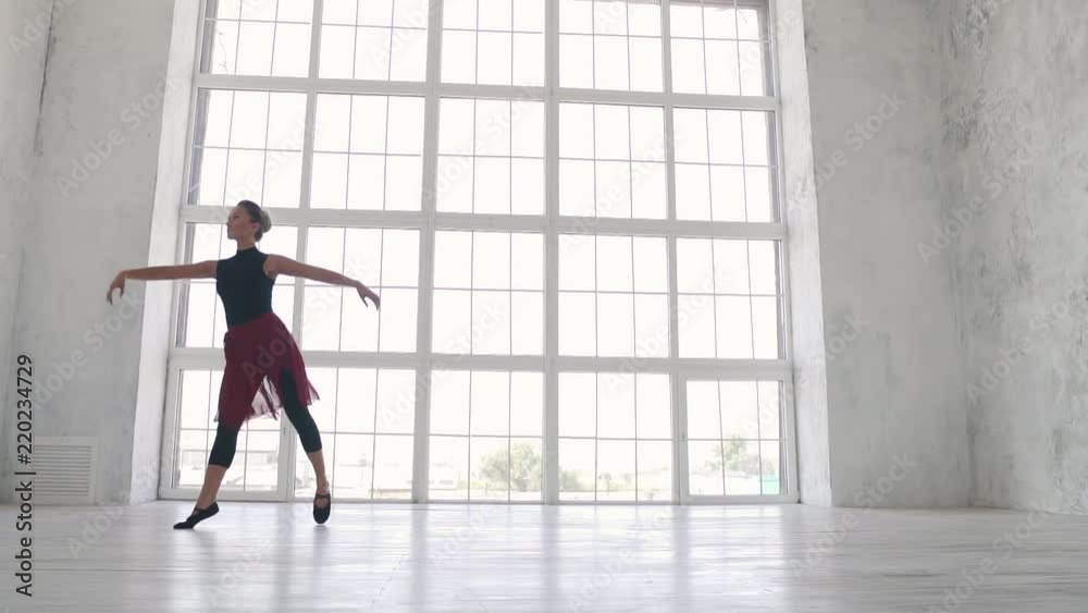 a ballerina in a black bodysuit and a red skirt dances a classic ballet in the Studio against a large light window. slow motion a ballerina in a black bodysuit and a red skirt dances a classic ballet in the Studio against a large light window. slow motion
