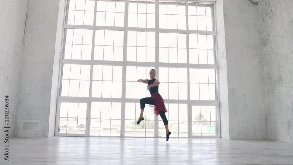 ballerina in a black bodysuit and a red skirt jumps high with a twine in the air in the Studio against a large light window. slow motion ballerina in a black bodysuit and a red skirt jumps high with a twine in the air in the Studio against a large light window. slow motion