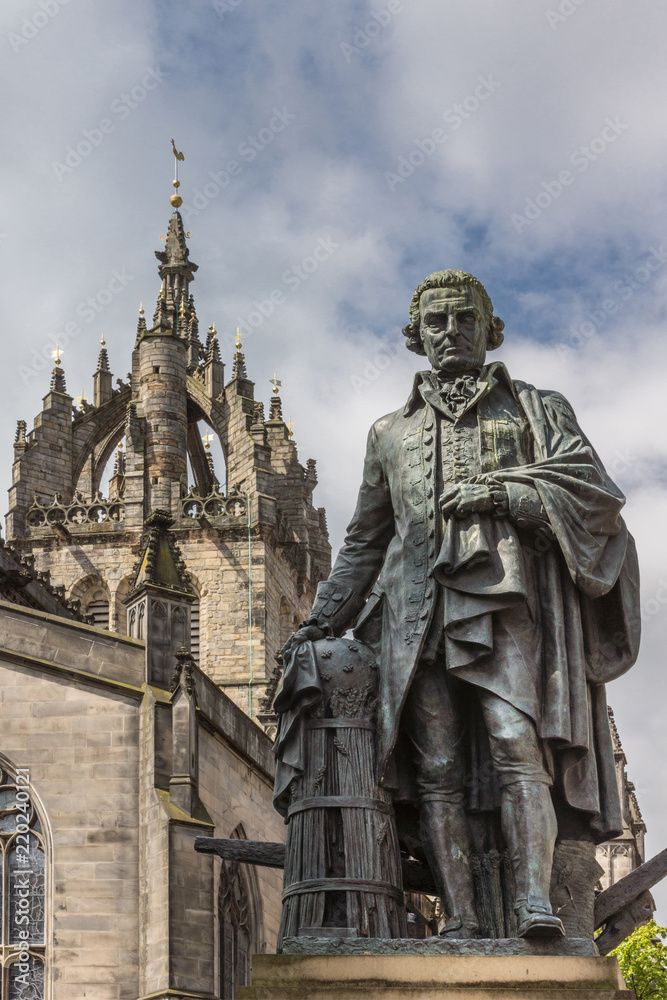 Edinburgh, Scotland, UK - June 14, 2012: Adam Smith bronze statue on market square in front of brown stone Saint Gilles Cathedral under blue sky with clouds. 