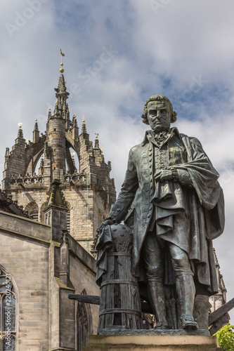 Edinburgh, Scotland, UK - June 14, 2012: Adam Smith bronze statue on market square in front of brown stone Saint Gilles Cathedral under blue sky with clouds. 