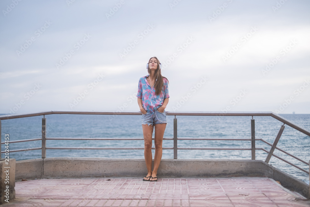 beautiful young lady caucasian woman standing at the beach on the road listening music with headphone and enjoying the leisure activity. vacation and lifestyle concept for nice people