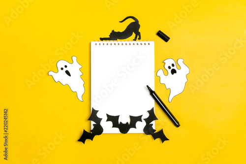 Crafts for the concept celebration of Halloween - figures of a black bat,cat,ghost cut from paper from stencil around a white notepad, felt-tip pen on yellow background Place for text Mock up Top view © bmarya83