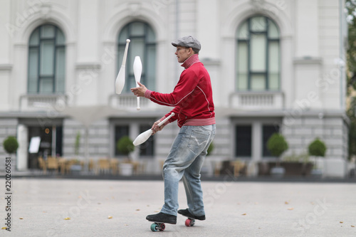 A man in a cap juggles with clubs, standing on small wheels on the street of a European city