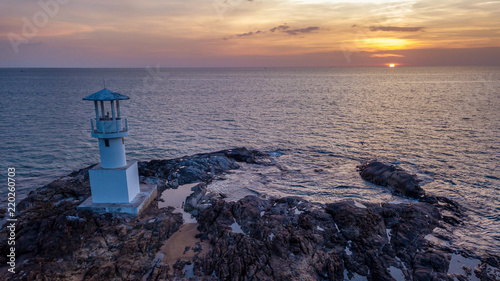 Drone view of a beautiful tropical sunset beind a small, white lighthouse located on rocks next to the ocean photo