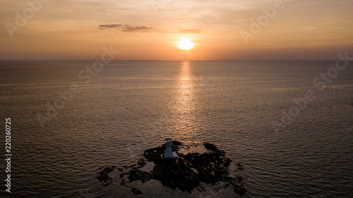 Drone view of a beautiful tropical sunset beind a small, white lighthouse located on rocks next to the ocean photo