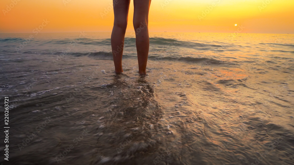 Fototapeta premium Woman feet splashed by sea waves on beach sunset. Calm serene relaxing scene of ocean water splashing on feet on beautiful beach