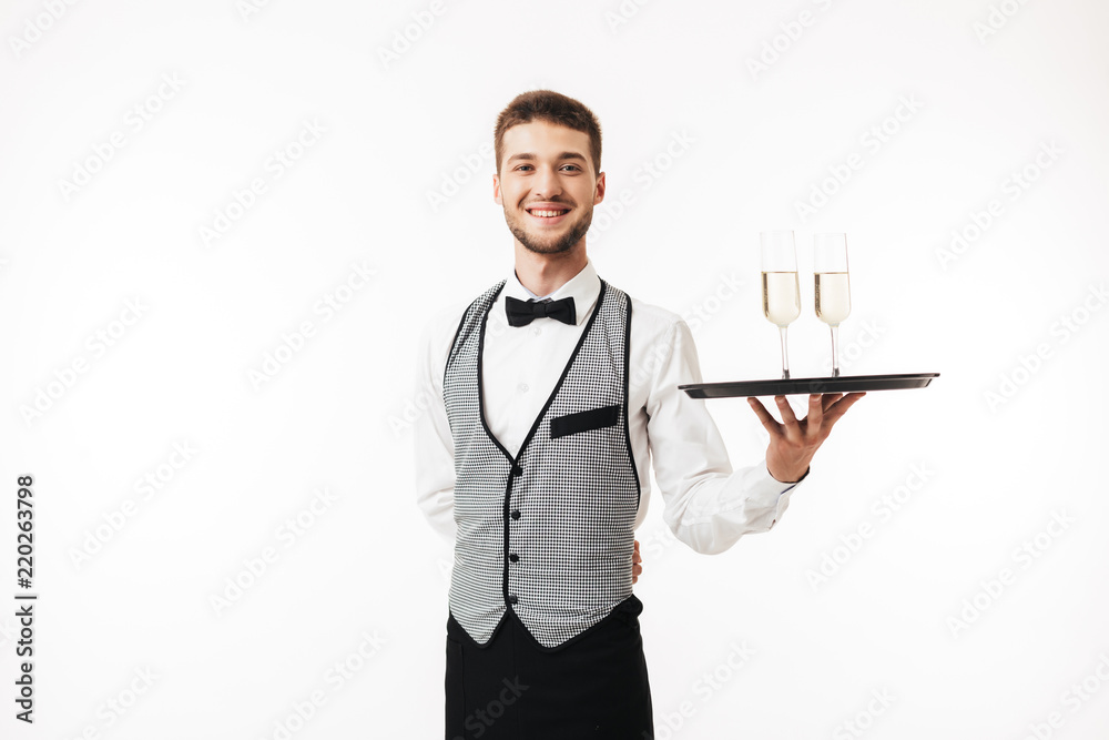 Joyful waiter in uniform happily looking in camera holding tray with glasses over white background isolated Joyful waiter in uniform happily looking in camera holding tray with glasses over white background isolated