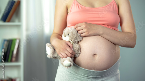 Dreamy woman with pregnant belly hugging toy bear tenderly, surrogate mother photo