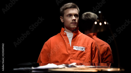 Canvas-taulu Handcuffed maniac in orange prison jumpsuit looking around in interrogation room