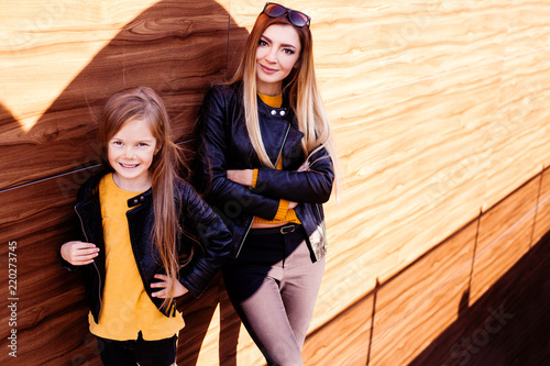 Smiling happy and looking sharp mother and daughter wearing black leather jacket on the stone background. Street fashion. City style. Kids fashion. Family look. Autumn. 