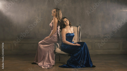 Two girls singer sit on a chair and sing. 4k longshot. Blue and pink evening luxurious dresses. Blond and Brunette woman.