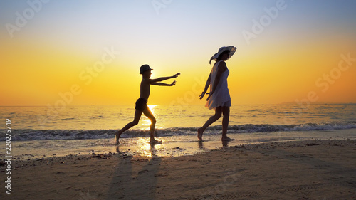 Family on summer vacation. Mother in white dress and son wearing hat walk on an epmty beach at vibrant sunset