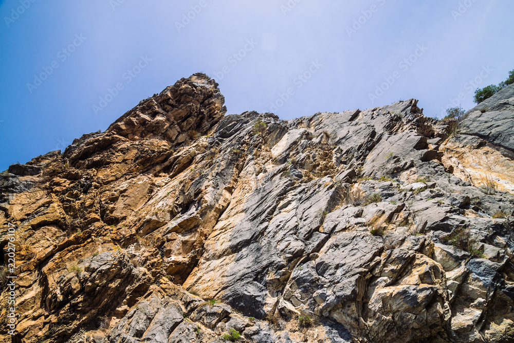 Big mountain cliff under cloudy sky close-up. Beautiful rocky gray textured background with vegetation. Majestic nature.
