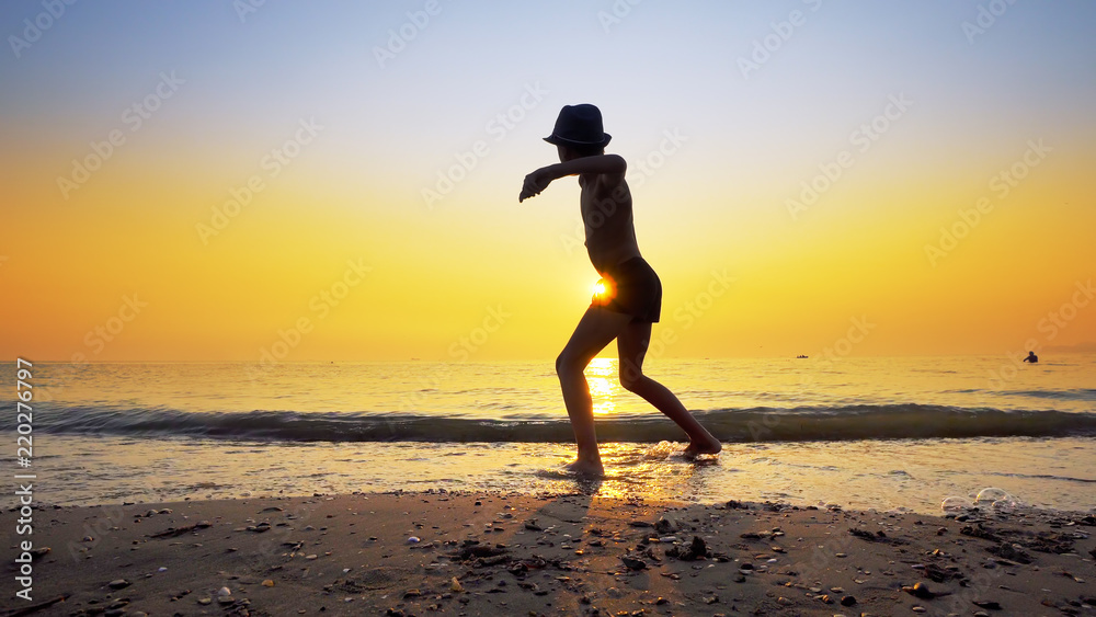 Young boy with hat collecting peebles on beach and throw stone skipping game on sea sunset water surface, small flattened rock bouncing off water surface across body of water many times